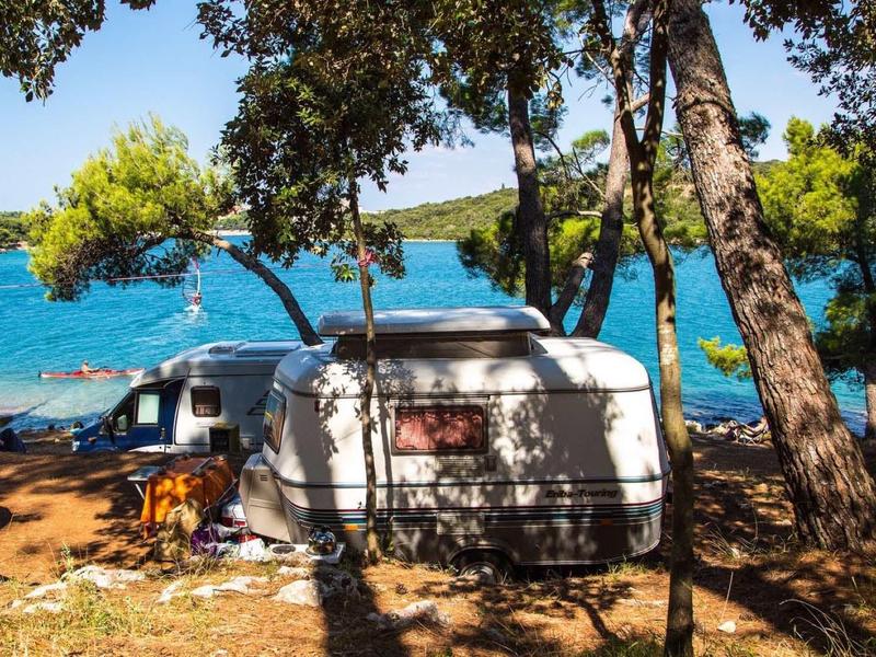 Wohnwagen steht im Schatten von Bäumen an einem klaren blauen See mit mehreren Booten.