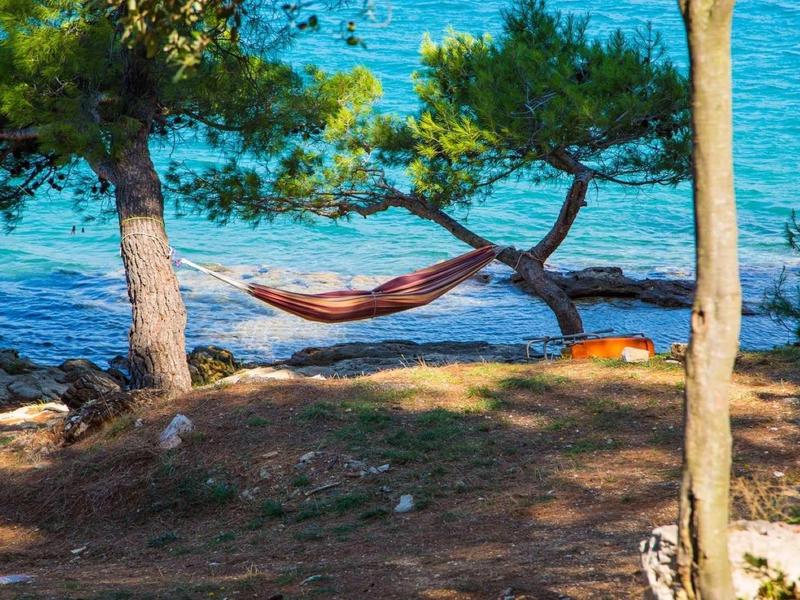 Person liegt in Hängematte zwischen Bäumen am Ufer mit blauem Meer im Hintergrund.