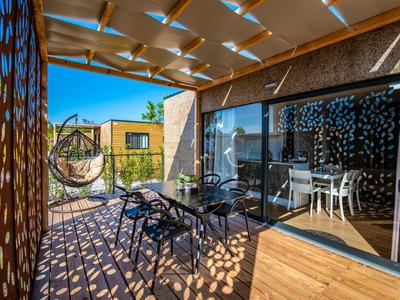 Otwarty taras z stołem, krzesłami i wiszącym fotelem, zacieniony przez pergolę z wzorzystym światłem.