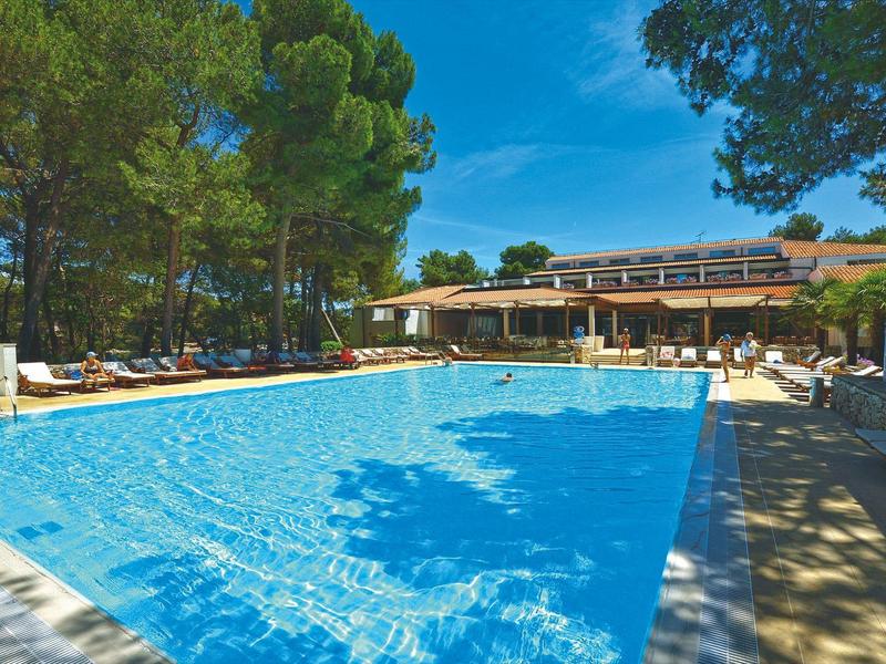 Duży basen na zewnątrz z parasolami i leżakami pod drzewami obok budynku hotelu.