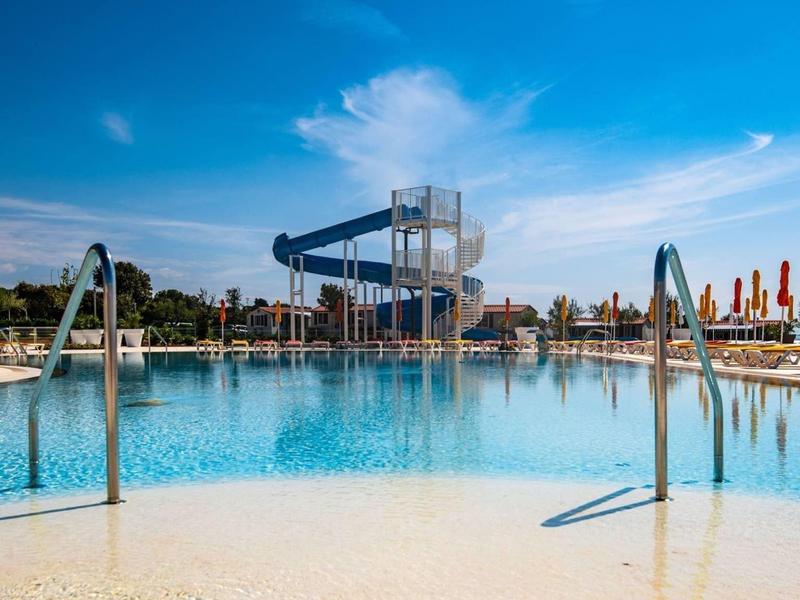 Przejrzysty basen z zjeżdżalnią wodną i parasolami pod błękitnym niebem.
