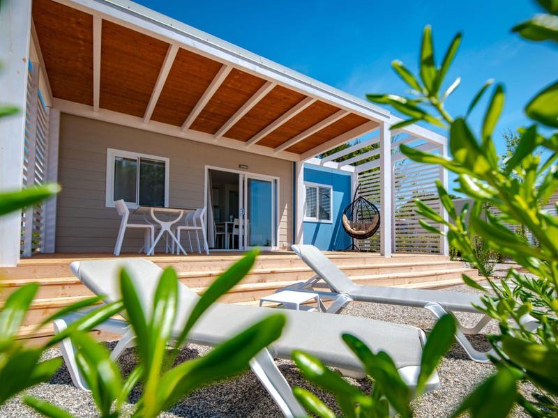 Nowoczesne patio z drewnianym tarasem, leżakiem, zestawem stołowym i przesuwnymi drzwiami pod błękitnym niebem.