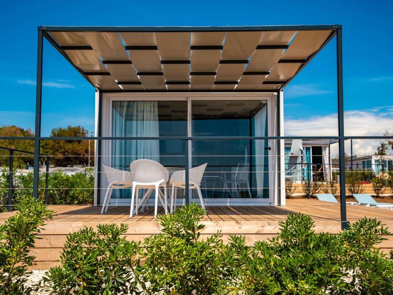 Taras nowoczesnej willi z stołem i krzesłami pod pergolą, otoczony zielenią.