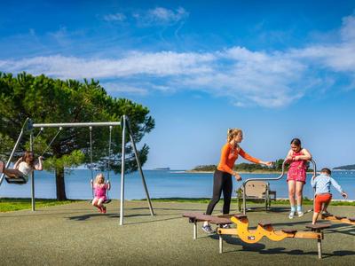 Kinder spielen auf einem Spielplatz mit Meerblick, blauem Himmel und Bäumen im Hintergrund.