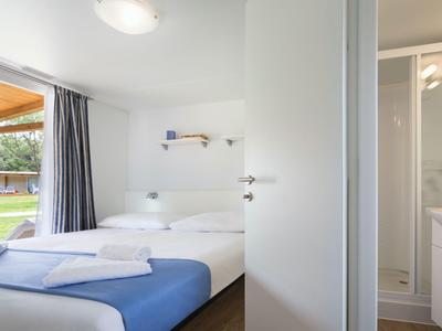 Helles Hotelzimmer mit Doppelbett, blauem Bettläufer und angrenzendem Bad mit weißer Einrichtung.