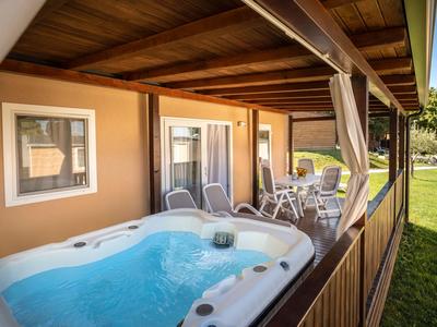 Überdachte Terrasse mit Whirlpool und Sitzbereich neben einem Ferienhaus mit Blick auf eine grüne Wiese.