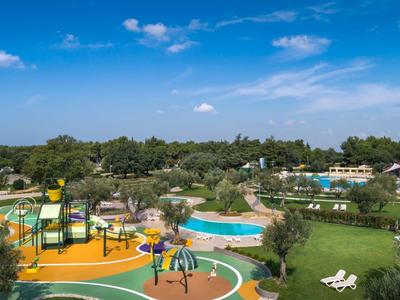 Blick auf einen Spielplatz und mehrere Pools in einer weitläufigen Hotelanlage unter blauem Himmel.
