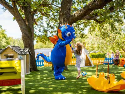 Kind im weißen Kleid spielt mit einem blauen Drachenmaskottchen im sonnigen Garten eines Hotels.