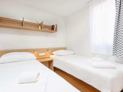 Helles Hotelzimmer mit zwei Einzelbetten, weißen Wänden und einem Holzregal über den Betten.