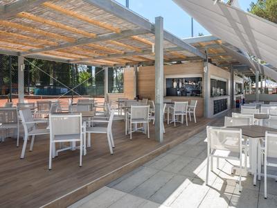 Offener Außenbereich eines modernen Cafés mit weißen Stühlen und Tischen auf Holz- und Fliesenboden unter Schattendach.