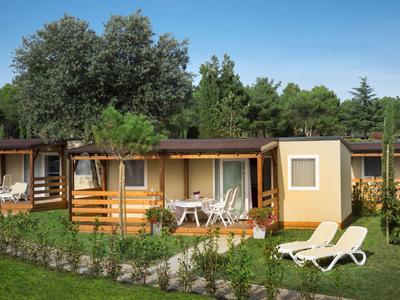 Casas de vacaciones modernas con terrazas de madera, árboles y tumbonas sobre césped verde.