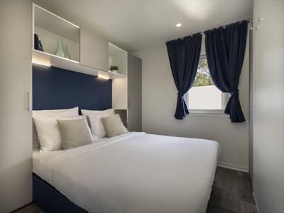 Dormitorio moderno con cama doble, paredes blancas y cortinas azules en ventana pequeña.