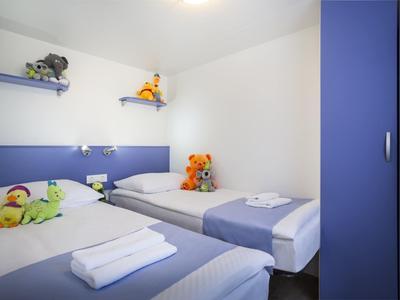 Pequeña habitación de hotel con dos camas individuales, muebles azules y peluches en las estanterías.