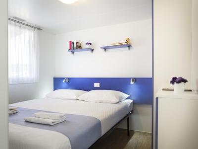 Habitación doble luminosa con paredes blancas, cabecera azul y dos estantes en la pared.