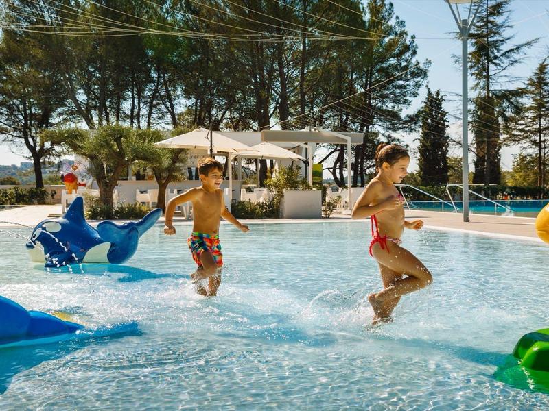 Niños juegan en piscina poco profunda con juguetes inflables coloridos bajo árboles.