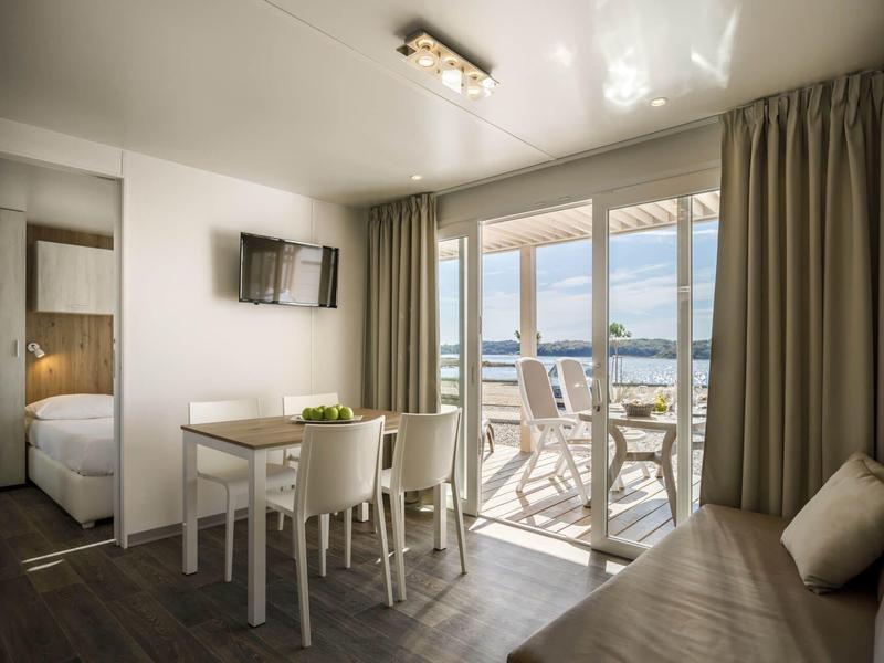 Habitación de hotel luminosa con mesa de comedor, TV, sofá y balcón con vista al mar.