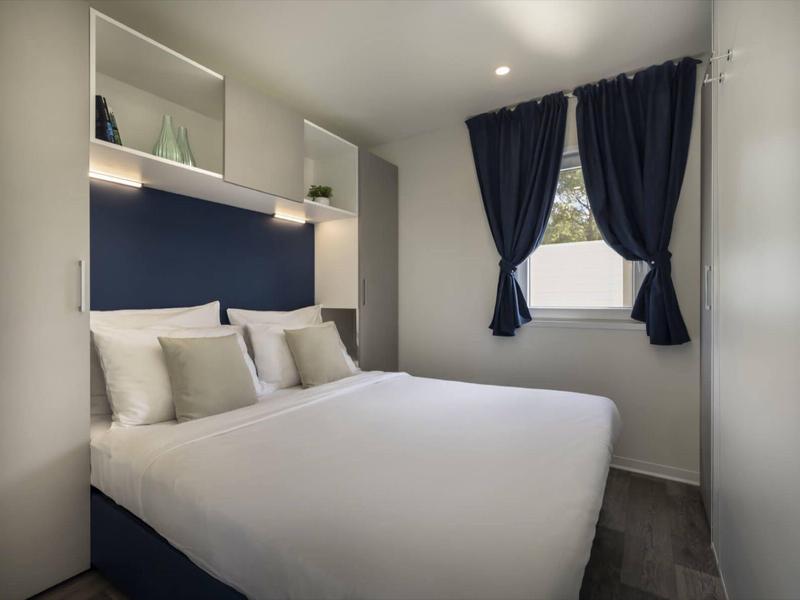 Dormitorio moderno con cama doble, paredes blancas y cortinas azules en ventana pequeña.