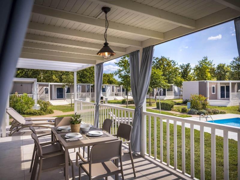 Terraza cubierta con mesa de comedor y vista a la piscina y casas modernas en entorno verde.