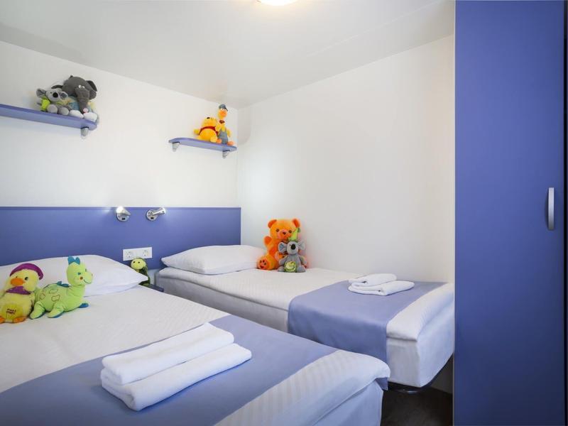 Pequeña habitación de hotel con dos camas individuales, muebles azules y peluches en las estanterías.