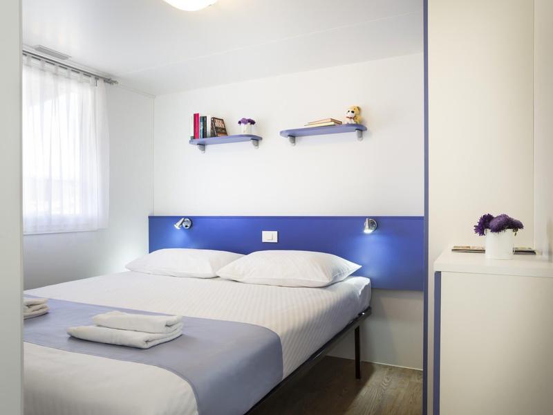 Habitación doble luminosa con paredes blancas, cabecera azul y dos estantes en la pared.