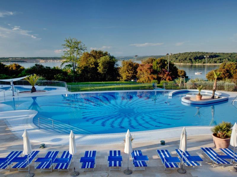 Buitenzwembad met blauwe ligstoelen met uitzicht op een meer en bomen onder een heldere lucht.