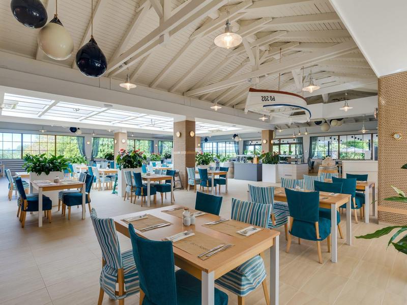 Ruime hotel eetzaal met houten tafels, blauwe stoelen en grote ramen die natuurlijk licht binnenlaten.