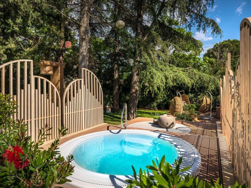 Kleine buiten jacuzzi omgeven door bomen en tuinplanten onder een heldere blauwe lucht.