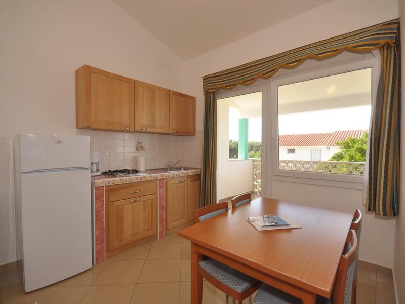 Malý kuchyňský kout s dřevěnými skříňkami, lednicí, stolem a židlemi u otevřeného okna.