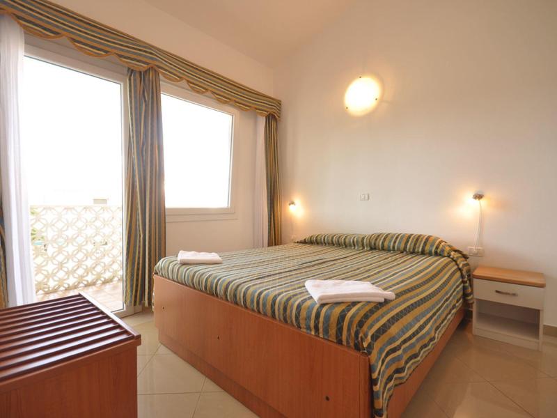 Světý hotelový pokoj s manželskou postelí, balkonem a jednoduchým dřevěným nábytkem.