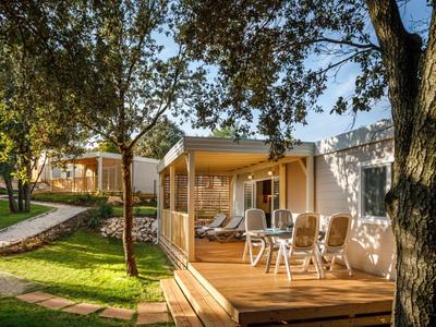 Holzterrasse mit weißem Tisch und Stühlen vor beigefarbenem Mobilheim, umgeben von Bäumen und Gras.