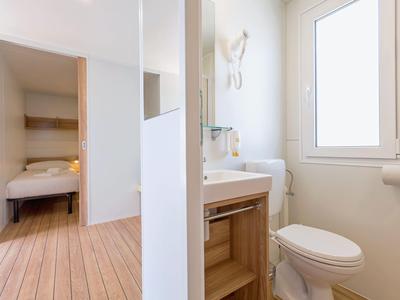 Baño compacto de hotel con inodoro, lavabo y vista al dormitorio contiguo.