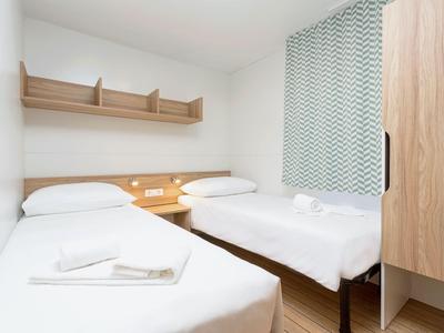 Habitación de hotel sencilla con dos camas individuales, estantes de madera y cortinas claras.