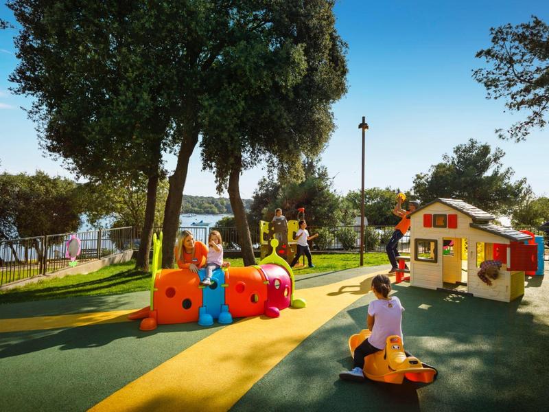 Bambini che giocano in un parco giochi colorato con scivoli e una casetta all'aperto con il sole.