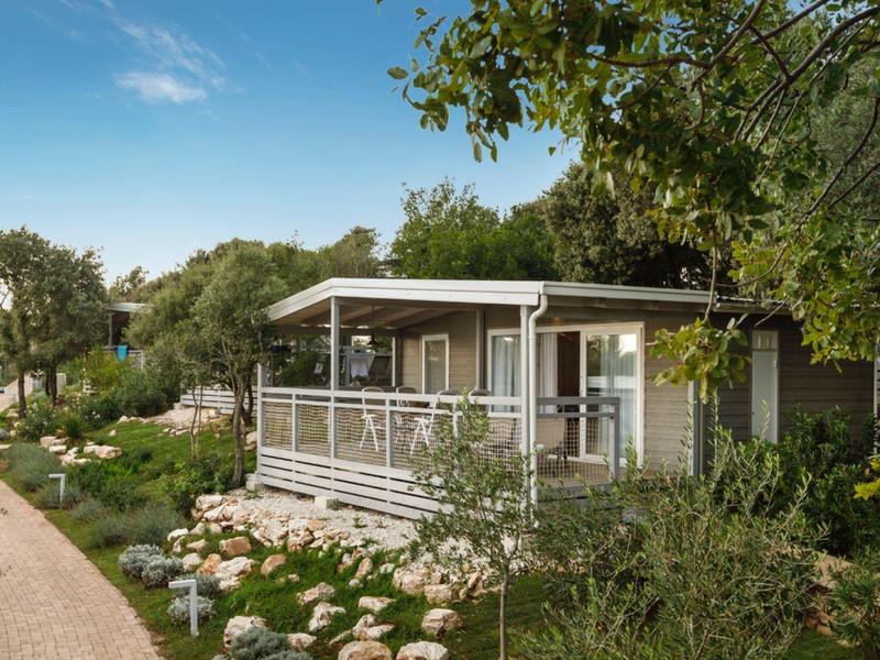 Casa vacanze moderna con terrazza, circondata da giardino e alberi sotto un cielo azzurro.