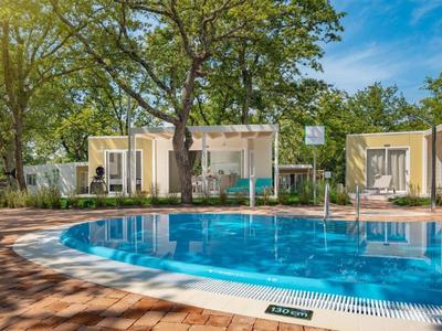 Runder Außenpool mit klarem Wasser, umgeben von braunem Pflaster und kleinen weißen Gebäuden.