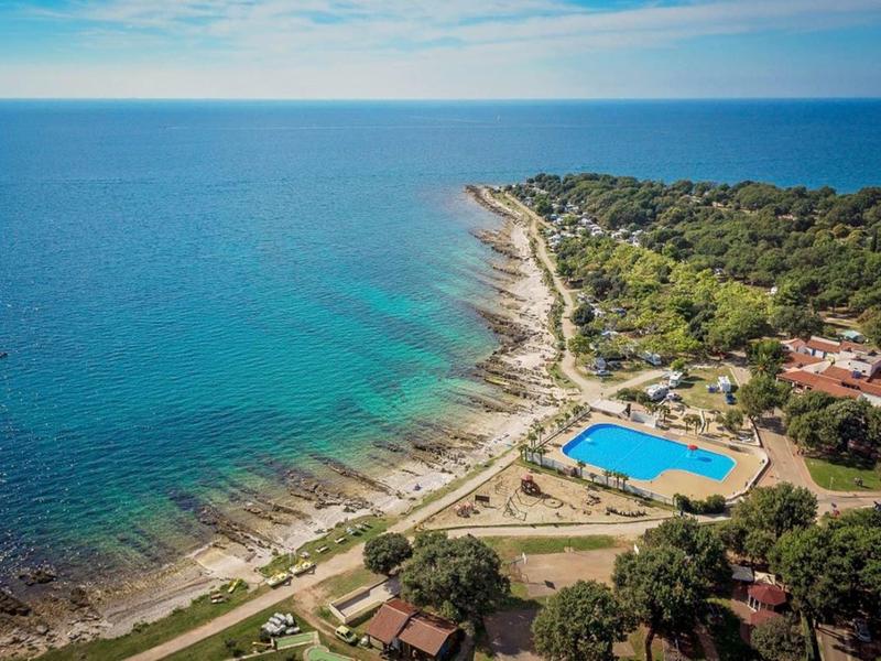 Küstenlinie mit klarem blauem Meer, Pool neben Waldgebiet und kleinen Gebäuden bei sonnigem Himmel.