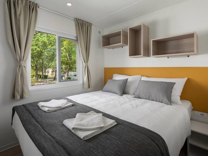 Helles Schlafzimmer mit großem Fenster, grauen Gardinen, weißem Bett und orangefarbener Wanddekoration.