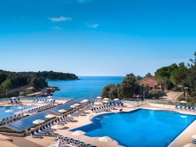 Großer blauer Pool mit Liegestühlen, umgeben von Bäumen und Blick aufs Meer bei klarem Himmel.