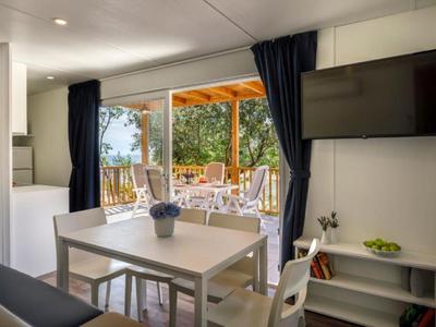 Salle à manger moderne avec table, chaises blanches et TV, ouverte sur balcon avec vue sur jardin.