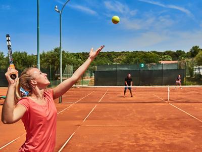 Une femme sert la balle sur un court de tennis en terre battue rouge par temps ensoleillé.