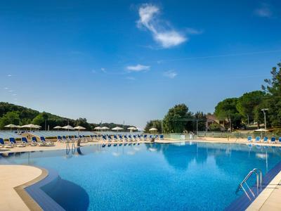 Großer Hotelpool mit blauer Wasseroberfläche und Liegestühlen unter sonnigem Himmel.