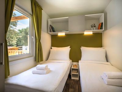Zweibettzimmer mit weißen Betten, grünen Vorhängen und moderner Holzdecke, Tageslicht durch Fenster.