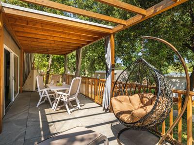 Balkon mit Holzdach und Möbeln neben einem hängenden Korbsessel mit Kissen, umgeben von Bäumen.