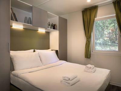 Schlafzimmer mit Doppelbett, frischer Bettwäsche, Fenster mit grünen Vorhängen und Regalen mit Dekoartikeln.