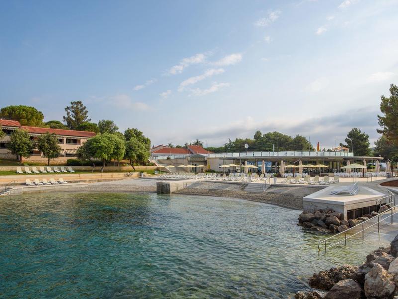 Clear water at a hotel bay with boat dock, sun loungers, and buildings in the background.