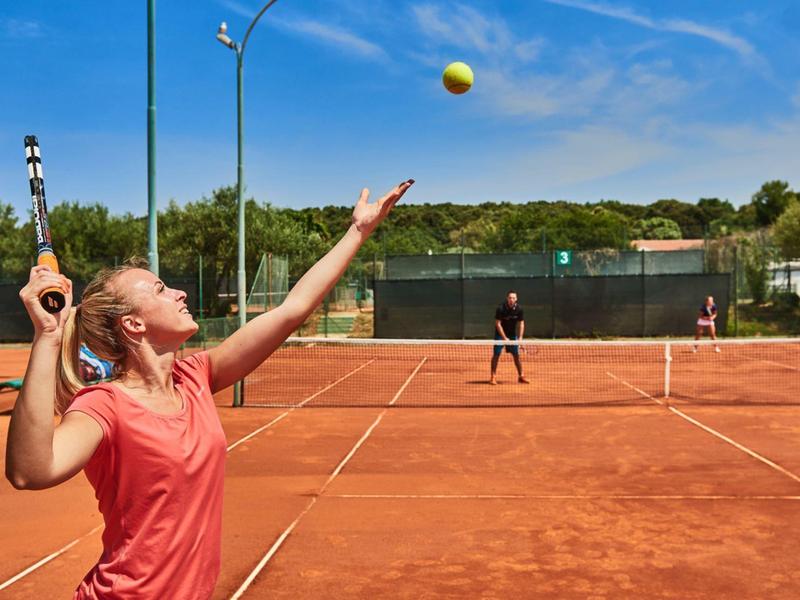 Woman in red sports top serving tennis ball on clay court in sunny weather.