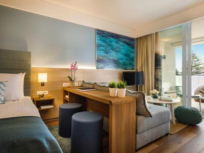 Modernes Hotelzimmer mit Bett, Sofa, Holztisch, blauem Hocker und Balkon mit Aussicht.