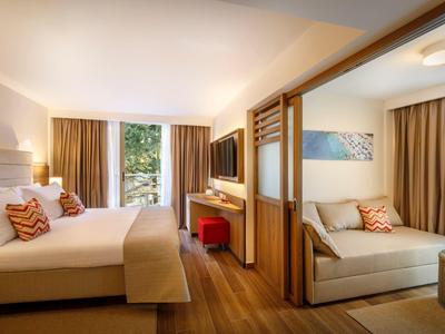 Modernes Hotelzimmer mit Doppelbett, Sofa, Holzboden und großen Fenstern mit Vorhängen.