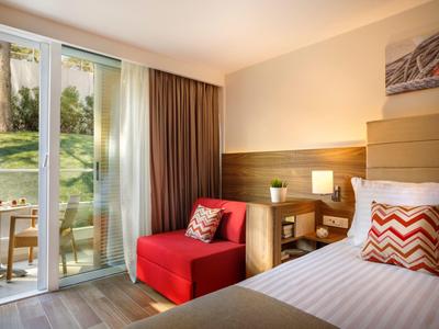 Hotelzimmer mit Einzelbett, rotem Sessel, Nachttischlampe und Balkon mit Sitzgelegenheit.