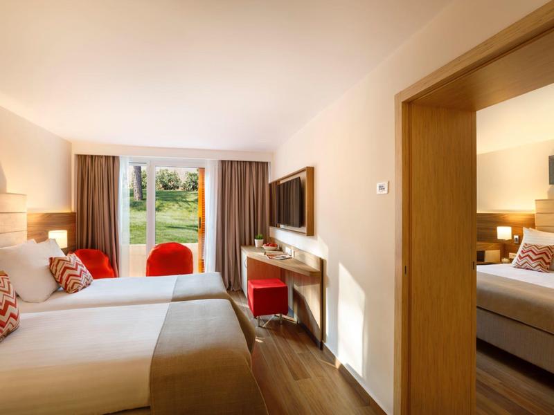 Helles Hotelzimmer mit Doppelbett, roten Stühlen, TV, Holzboden und Blick auf Garten durch Fenster.